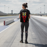 A woman stands on a racetrack wearing a black backpack with a large red, yellow, and white graphic design on the back. She is seen from behind, wearing a black t-shirt, ripped black jeans, and black boots.