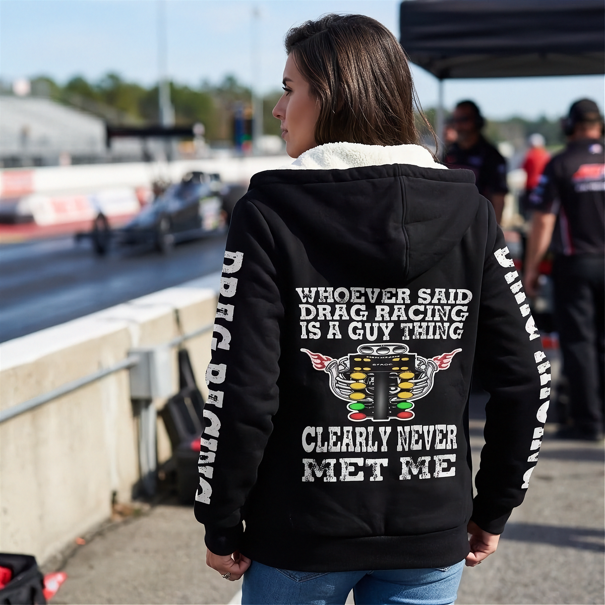 A woman modeling a black zip-up hoodie with a light beige fleece lining. The hoodie features a white graphic print on the back and white DRAG RACING on the sleeves.