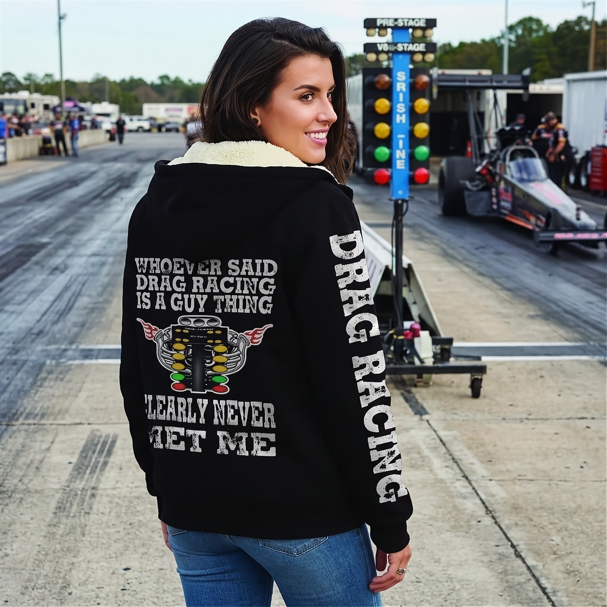 A woman modeling a black zip-up hoodie with a light beige fleece lining. The hoodie features a white graphic print on the back and white DRAG RACING on the sleeves.