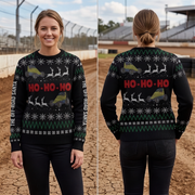 A woman modeling a black Christmas sweater in a split front-and-back view, standing outdoors on a dirt track. The sweater has a festive holiday pattern with green snowflakes, white reindeer, yellow off-road vehicles, and red 'HO-HO-HO' text.