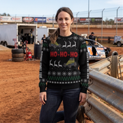A woman stands at an outdoor dirt track wearing a black Christmas sweater with red 'HO-HO-HO' lettering, white reindeer silhouettes, green zigzag stripes, and yellow off-road vehicle graphics. Racing trailers, tires, and a race car are visible in the background.