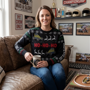 A woman sitting on a brown leather couch wearing a black Christmas sweater with green and red patterns, yellow and blue race cars, and 'HO-HO-HO' in red text. She is holding a ceramic mug in a decorated garage setting with racing memorabilia.