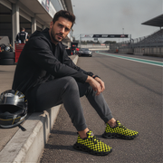 Man sitting on a race track wearing checkered shoes, with a helmet beside him.