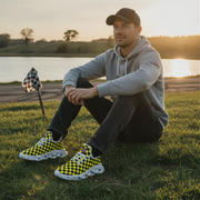 Man sitting on grass wearing checkered shoes with a sunset and lake in the background