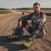 Man sitting on a dirt road wearing yellow checkered shoes, with another person on a motorcycle in the background.