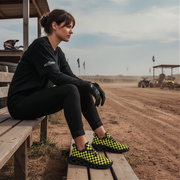 Woman sitting on a wooden bench at an outdoor event with vehicles and flags in the background.