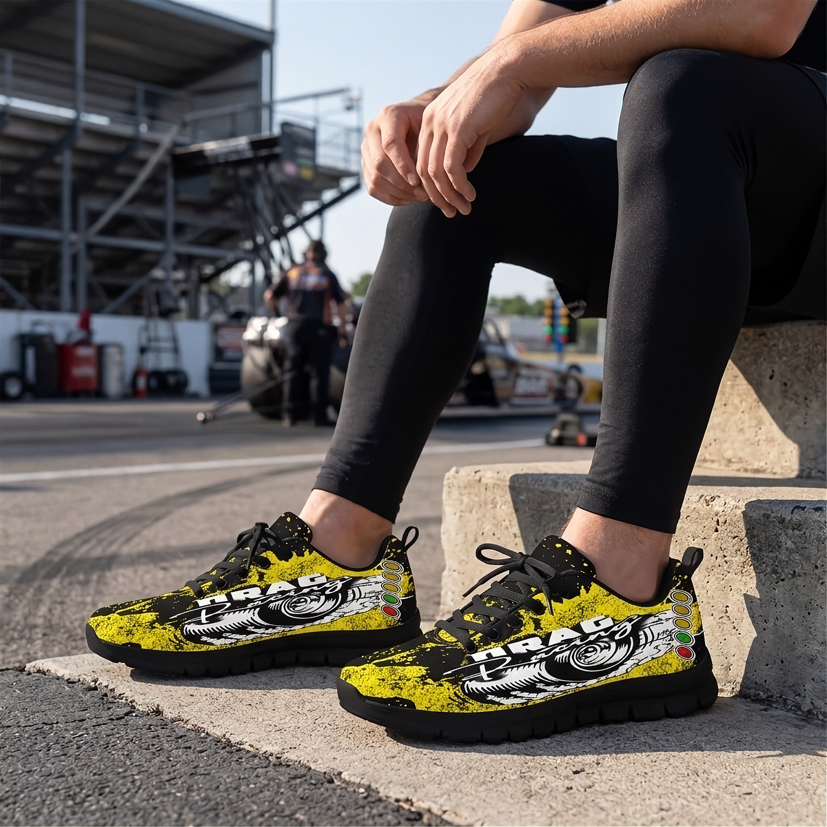 A person sitting on concrete at a racetrack wearing a pair of black, yellow, and white racing-style sneakers with a motorcycle graphic and the word 'DRAG RACING'.