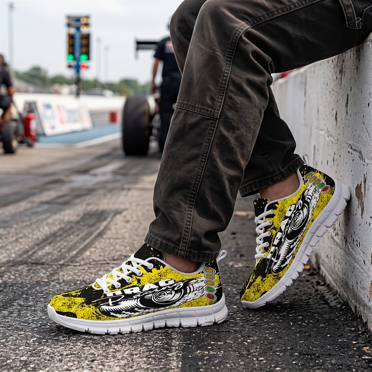 A person sitting on a concrete wall at a racetrack wearing a pair of low-top sneakers with a bold yellow, black, and white graphic design. The background shows blurred racetrack elements and another person in the distance.