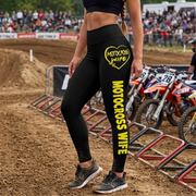Black high-waisted leggings with yellow 'MOTORCROSS WIFE' text and a heart graphic on the thigh, worn by a model on a dirt motocross track with race bikes in the background.