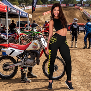 A woman stands on a dirt motocross track next to a red and white Honda dirt bike, wearing a black crop top and black leggings with yellow graphics. Other motocross bikes, riders, tents, and track signage are visible in the background.
