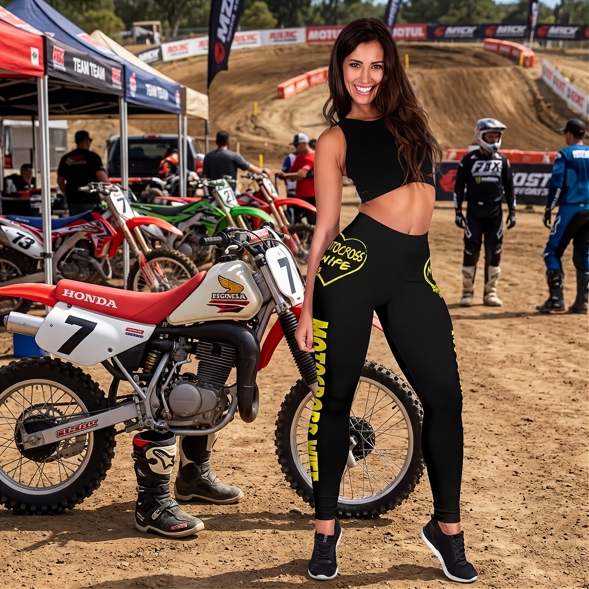 A woman stands on a dirt motocross track next to a red and white Honda dirt bike, wearing a black crop top and black leggings with yellow graphics. Other motocross bikes, riders, tents, and track signage are visible in the background.
