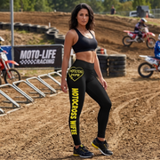 A woman stands on a dirt motocross track wearing black athletic leggings with yellow 'MOTOCROSS WIFE' text and a yellow heart logo on the thigh, along with a black sports bra and black sneakers. Dirt bikes and track barriers are visible in the background.