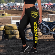 A person wearing black high-waisted leggings with bright yellow 'MOTOCROSS WIFE' text and a heart graphic, standing in a dirt motocross arena near stacked tires and a motorcycle.