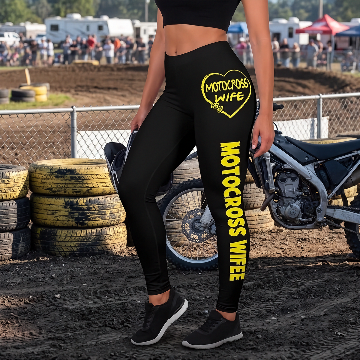 A person wearing black high-waisted leggings with bright yellow 'MOTOCROSS WIFE' text and a heart graphic, standing in a dirt motocross arena near stacked tires and a motorcycle.