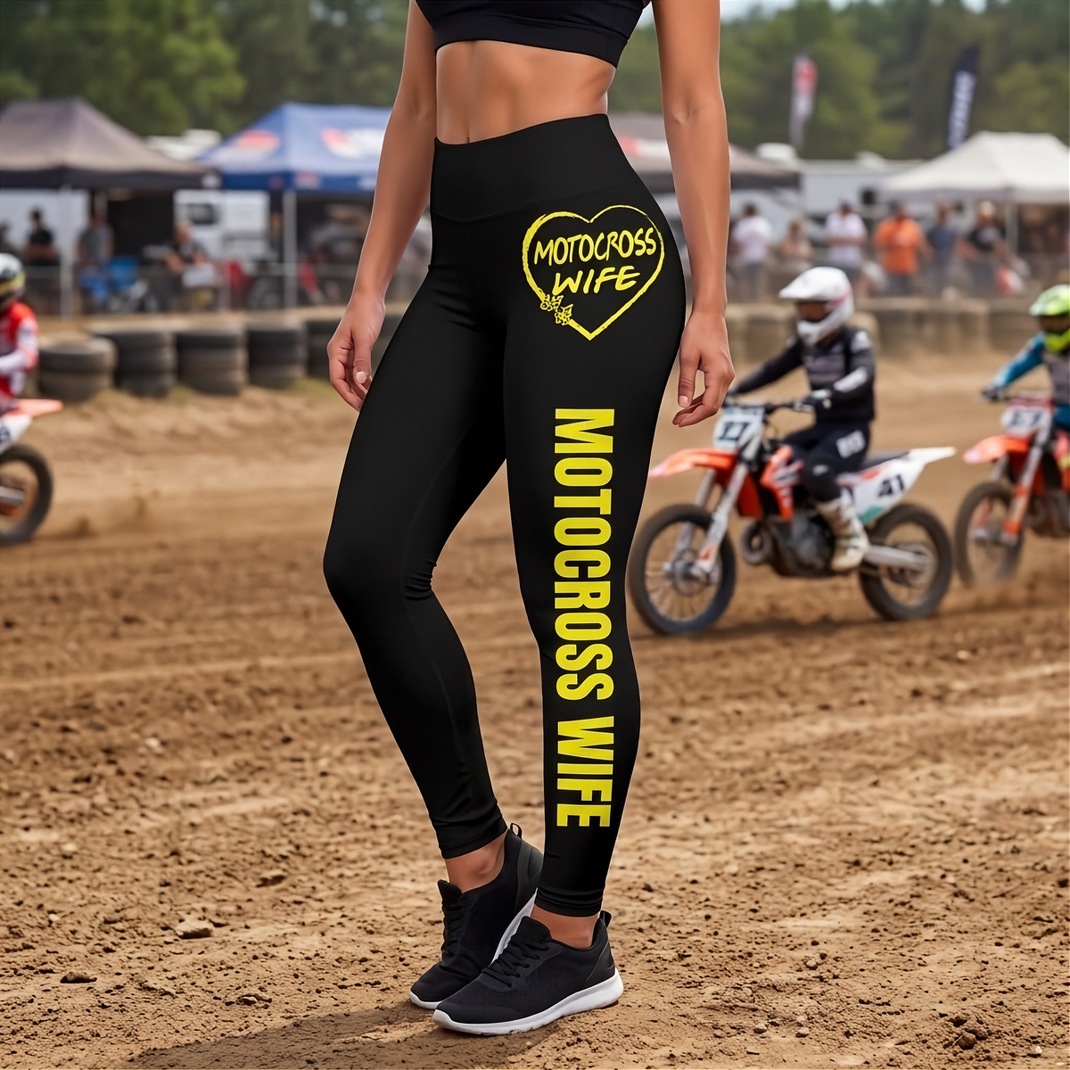 Black high-waisted leggings with yellow 'MOTOCROSS WIFE' text and a heart graphic on the thigh, worn by a model on a dirt motocross track with race bikes in the background.