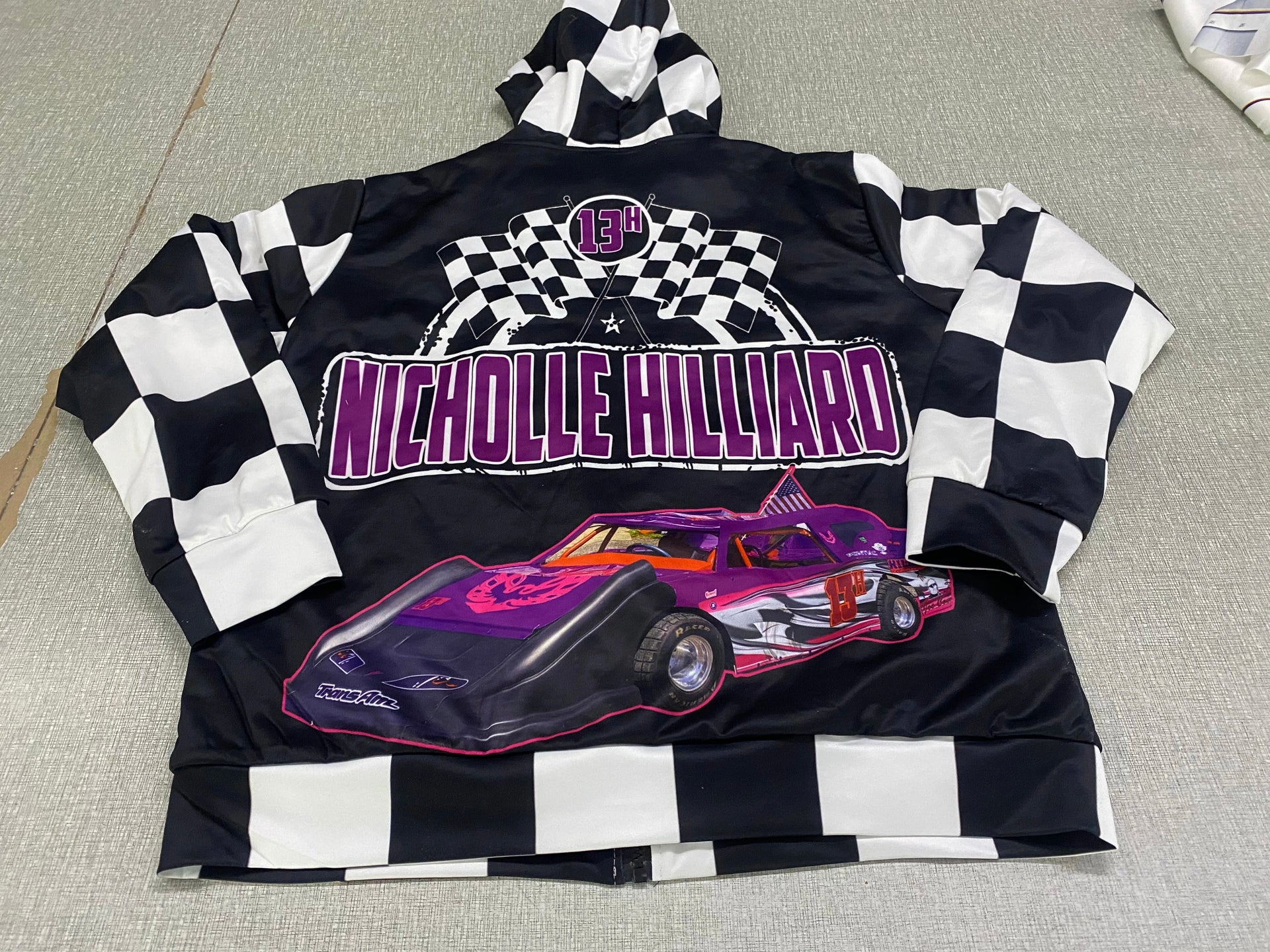 A personalized racing jacket displayed on a mannequin, featuring a large, high-resolution photo of a custom muscle car printed on the back panel, complemented by black and white checkered flag accents on the sleeves and a plush, cream-colored sherpa lining visible at the collar.