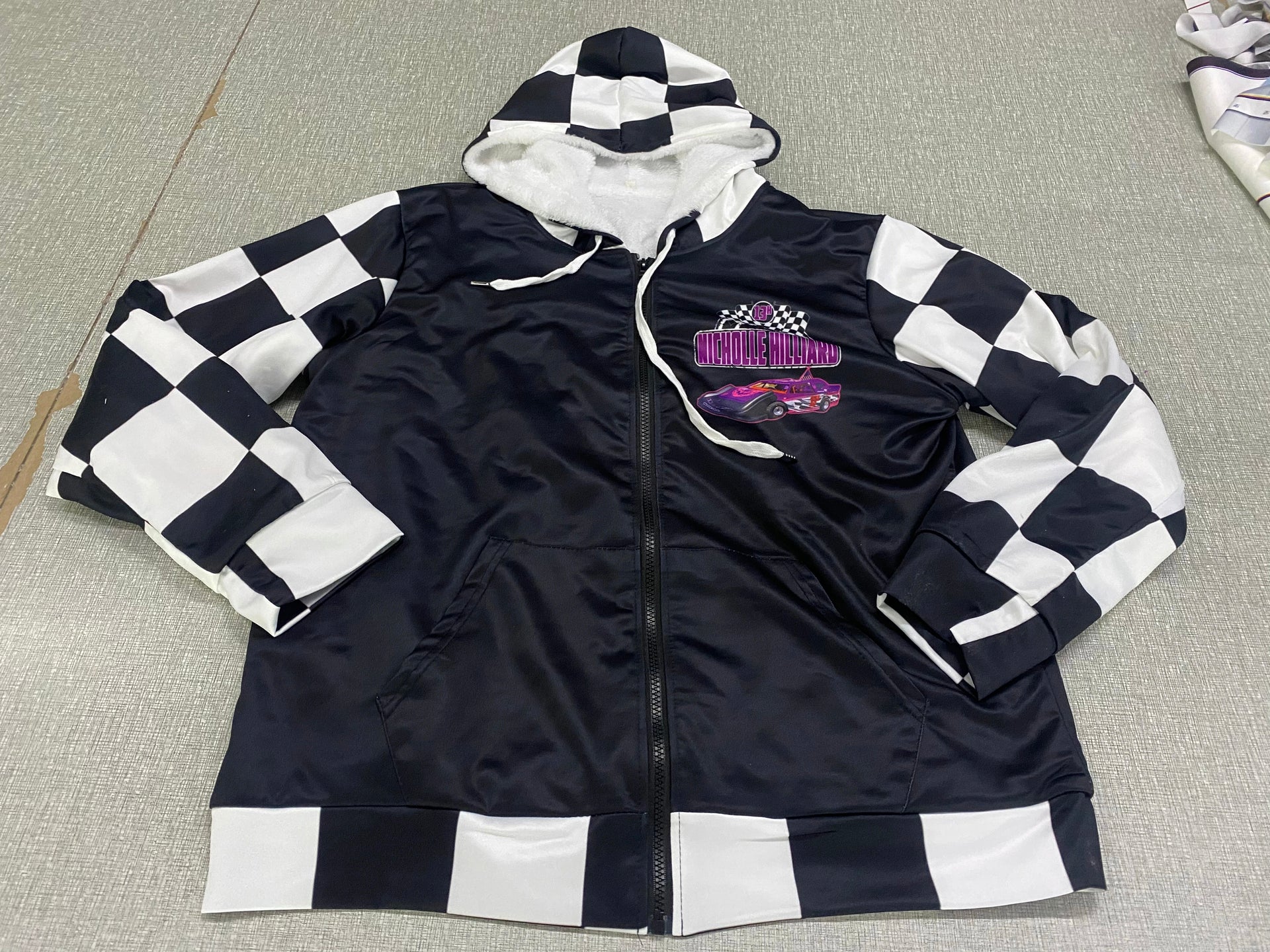 A personalized racing jacket displayed on a mannequin, featuring a large, high-resolution photo of a custom muscle car printed on the back panel, complemented by black and white checkered flag accents on the sleeves and a plush, cream-colored sherpa lining visible at the collar.