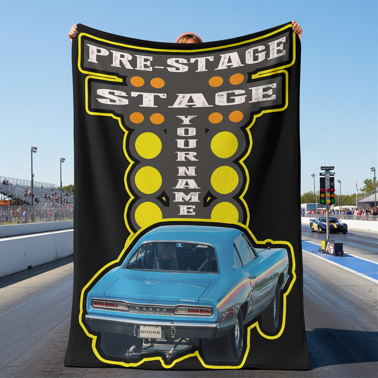 Person holding a large blanket with a blue car graphic and text at a drag race track.
