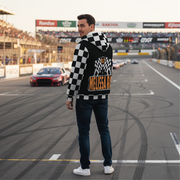 A race fan wearing a custom-printed sherpa-lined racing jacket while standing in the grandstands of an illuminated dirt track at night, with the race cars visible in the background.