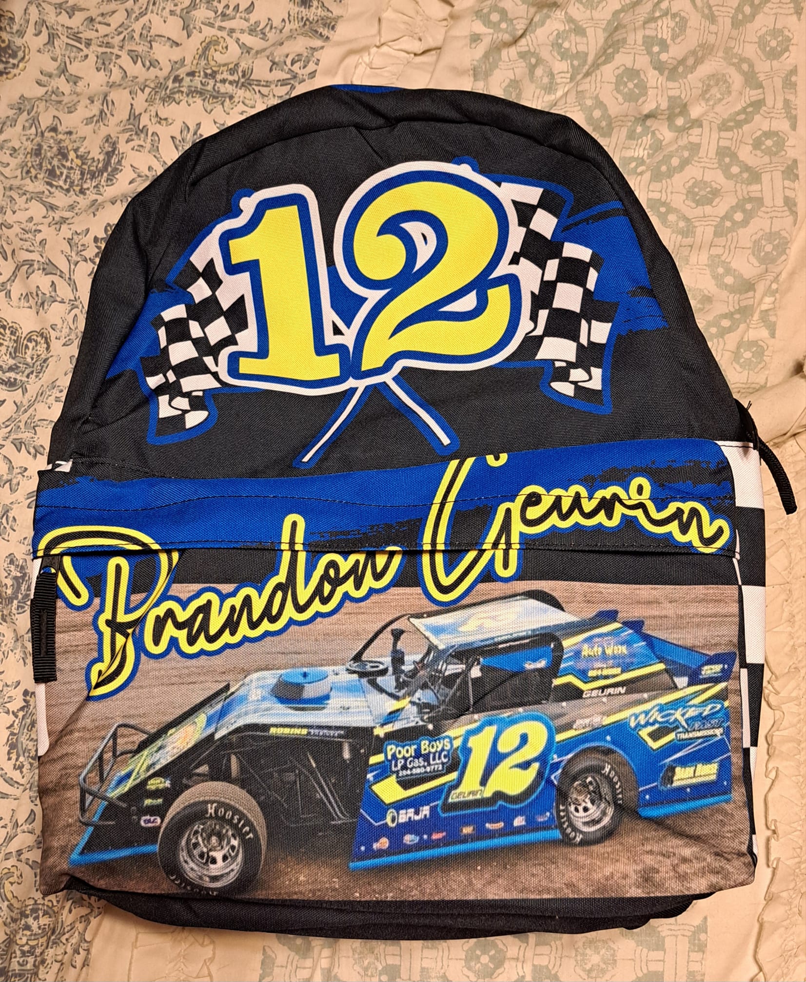 A checkered backpack with a racing theme, featuring a large main compartment and a front utility pocket. The backpack is custom-made with a blue and black checkered pattern and has a graphic of a racing car with the number 12 and the name 'Brandon Geurin'.