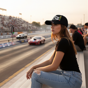 Woman sitting at a drag race track with cars on the side wearing a drag racing baseball cap