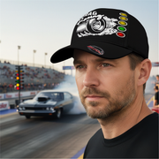 Man wearing a black cap with drag racing-themed design at a drag race track.