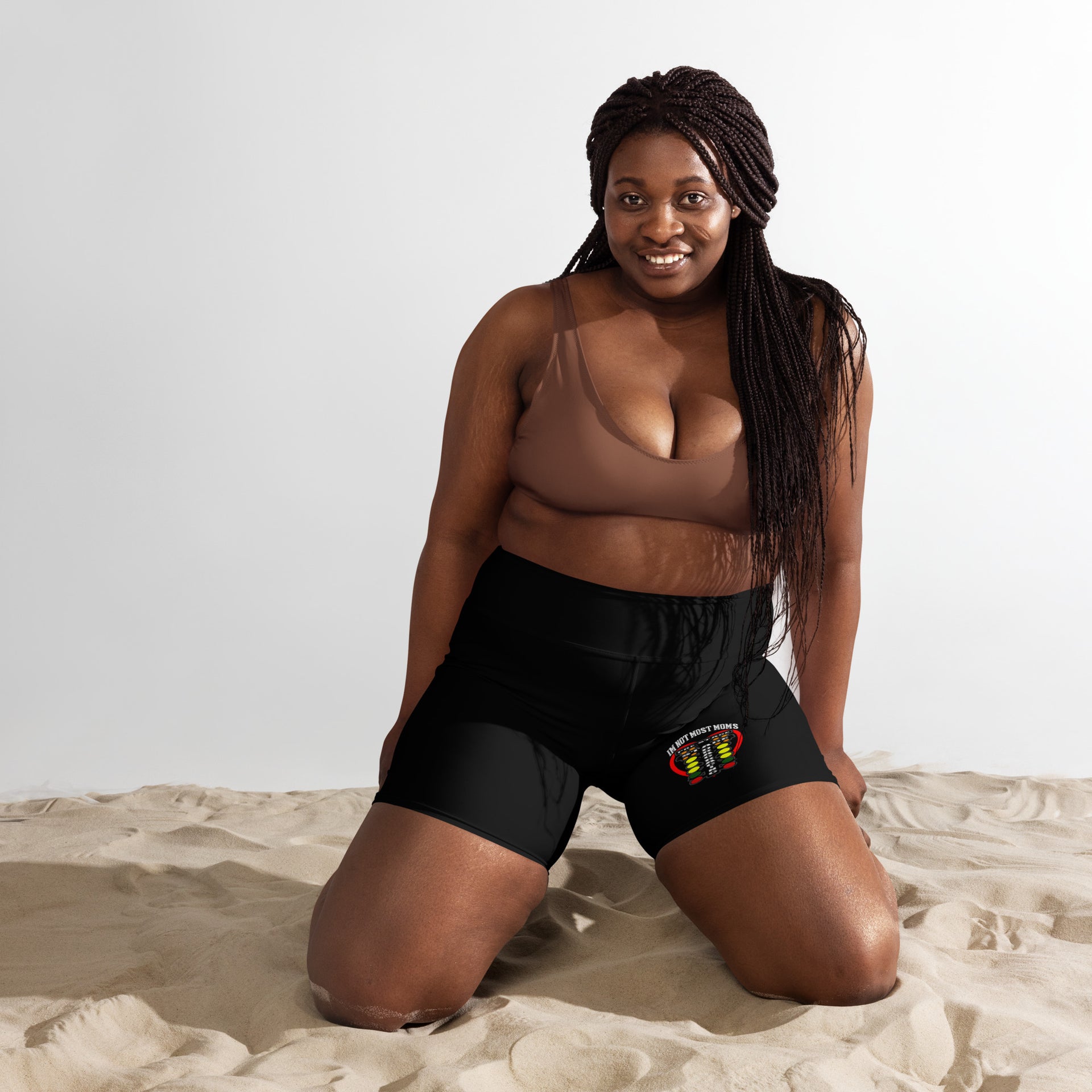 Woman sitting on sand wearing a brown top and black shorts with a white logo.
