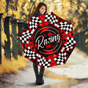 A woman holding a large open racing-themed umbrella in an outdoor autumn setting with trees and a wet path. The umbrella features a red and black center with the word 'Racing' in white script, surrounded by black-and-white checkered segments.