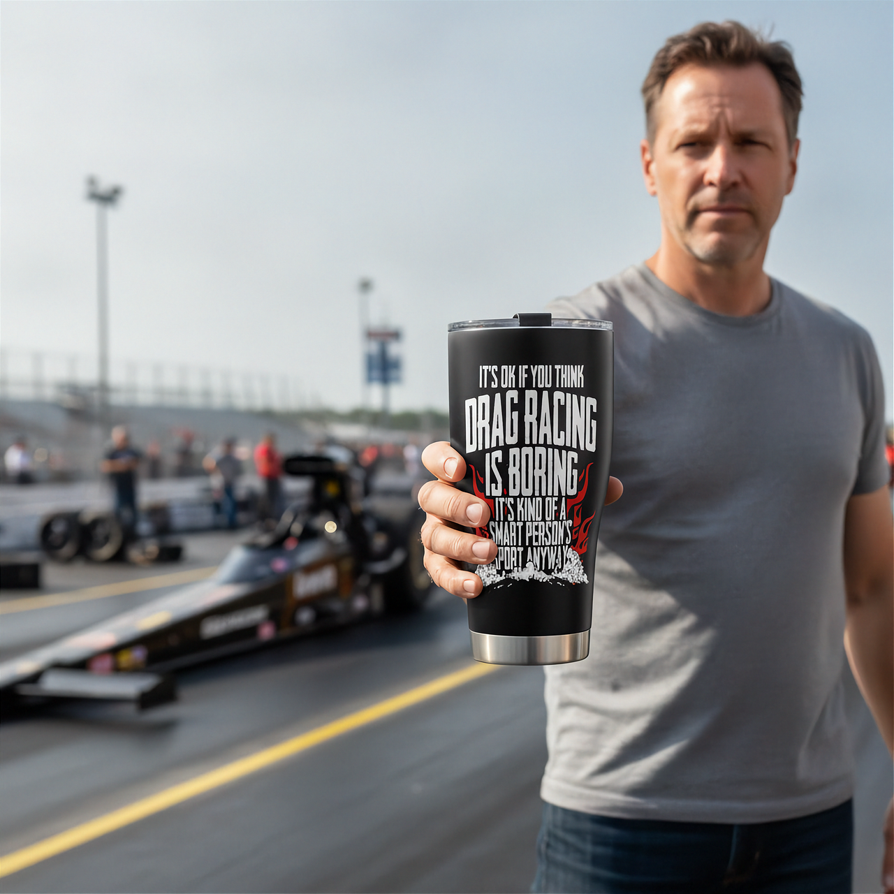 A man in a gray t-shirt stands on a drag racing track holding a black travel tumbler with a silver stainless steel rim. The tumbler features white text that reads 'IT'S OK IF YOU THINK DRAG RACING IS BORING.