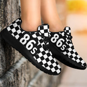 Black and white checkered sneakers with '86s' printed in white on the side, shown on a person's feet leaning against a stone wall.