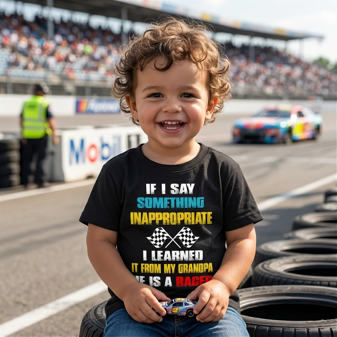 A smiling toddler wearing a funny "If I Say Something Inappropriate I Learned It From My Grandpa" racing t-shirt, sitting on a mechanic's creeper in a home garage alongside their grandfather who is holding a wrench and laughing.