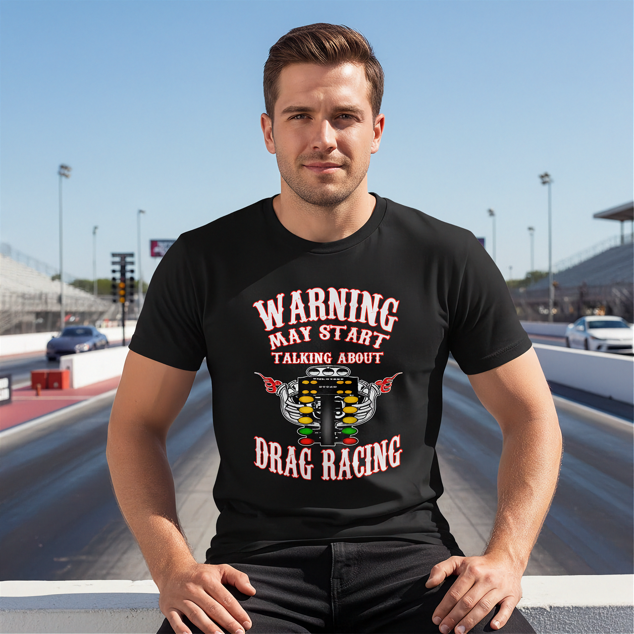 A smiling adult man wearing a black "Warning May Start Talking About Drag Racing" graphic hoodie while working on the open engine bay of a classic muscle car inside a well-lit garage workshop.
