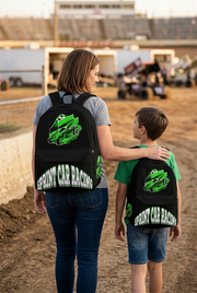 A young boy wearing a vivid green sprint car racing backpack, standing near a chain-link fence at a dirt oval track under bright stadium lights, watching winged sprint cars slide through a muddy corner.