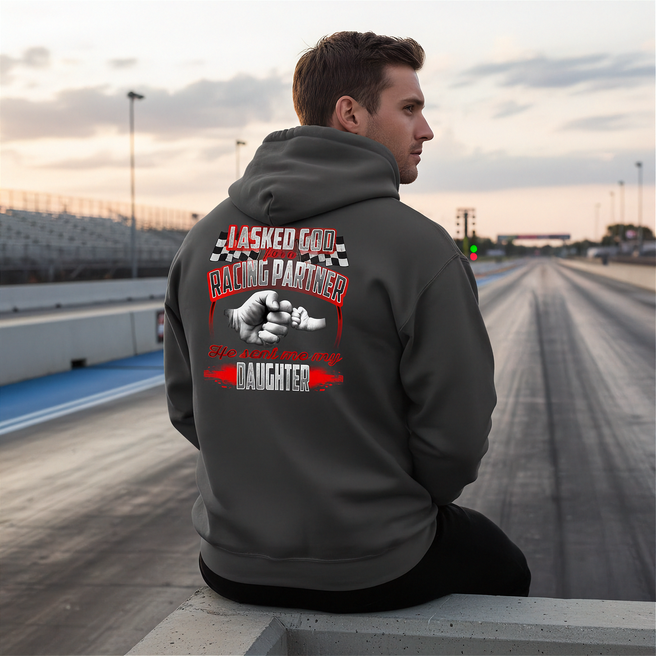 A proud father wearing the "I Asked God For A Racing Partner" graphic t-shirt standing in a paved race track pit lane, smiling beside his young daughter who is sitting on the tire of a custom quarter-mile junior dragster.