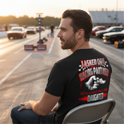 A proud father wearing the "I Asked God For A Racing Partner" graphic t-shirt standing in a paved race track pit lane, smiling beside his young daughter who is sitting on the tire of a custom quarter-mile junior dragster.