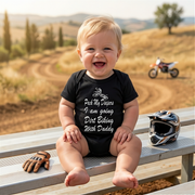 A proud father dressed in bright motocross gear holding his laughing baby, who is wearing the "Pack My Diapers" dirt biking bodysuit, standing next to a muddy off-road motorcycle on a sunny forest trail.