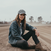 Woman sitting on a dirt track wearing a denim jacket, checkered cap, and brown boots.