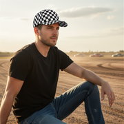 Man wearing a checkered cap and black shirt sitting on a dirt track with cars in the background.