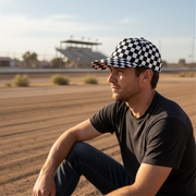 Man wearing a checkered cap and black t-shirt sitting on a dirt track.