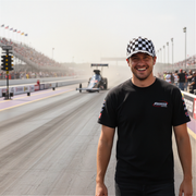 Man in a checkered cap and black t-shirt standing on a race track with racing cars in the background.
