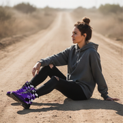Woman sitting on a dirt road wearing a gray hoodie and black pants with purple checkered boots.