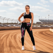 Woman in athletic wear standing on a dirt track with cars and a cloudy sky in the background wearing a racing girl leggings