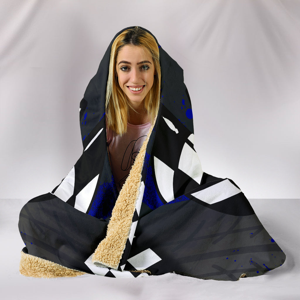 A woman sitting wearing a hooded blanket with a drag racing theme. The garment features a black-and-white checkered pattern, blue splatter accents, and a central vertical graphic with the words 'PRE-STAGE', 'STAGE', and 'DRAG RACING' over a red, yellow, green, and orange light-tower motif.