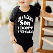 A toddler wearing a black long sleeve baby bodysuit with the text 'I'm a RACER'S SON I DON'T KEEP CALM' printed on the front.