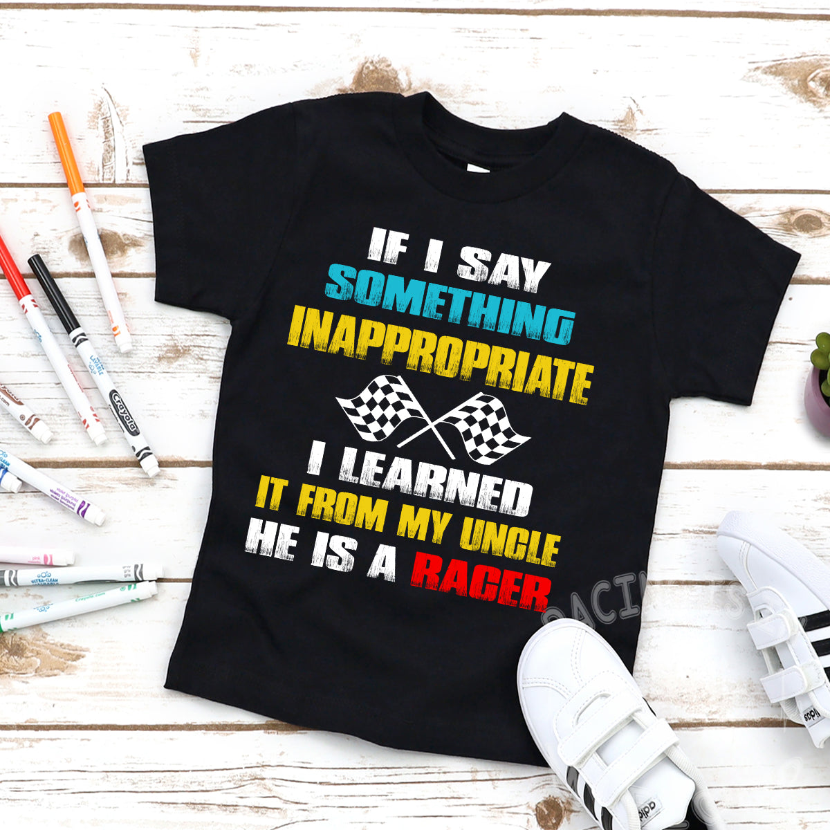 Black youth t-shirt with text 'If I say something inappropriate I learned it from my uncle he is a Rager' printed on it, presented on a wooden surface with markers and other crafting materials around it.