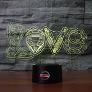 Love Motocross 3D LED Lamp showing a dirt bike rider jumping through a heart-shaped silhouette, glowing in a vibrant pink on a modern black base.