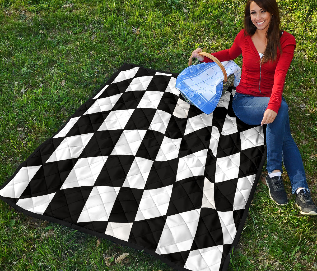 A woman sitting on a black and white checkered quilted blanket in a grassy field under a blue sky with clouds.