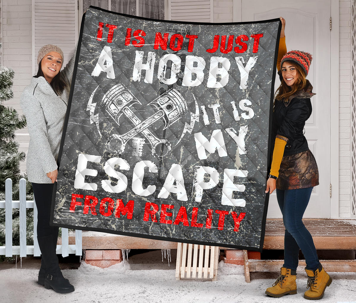Two women displaying the gray piston quilt outside a snowy porch, showing distressed red-and-white lettering and diamond stitching.
