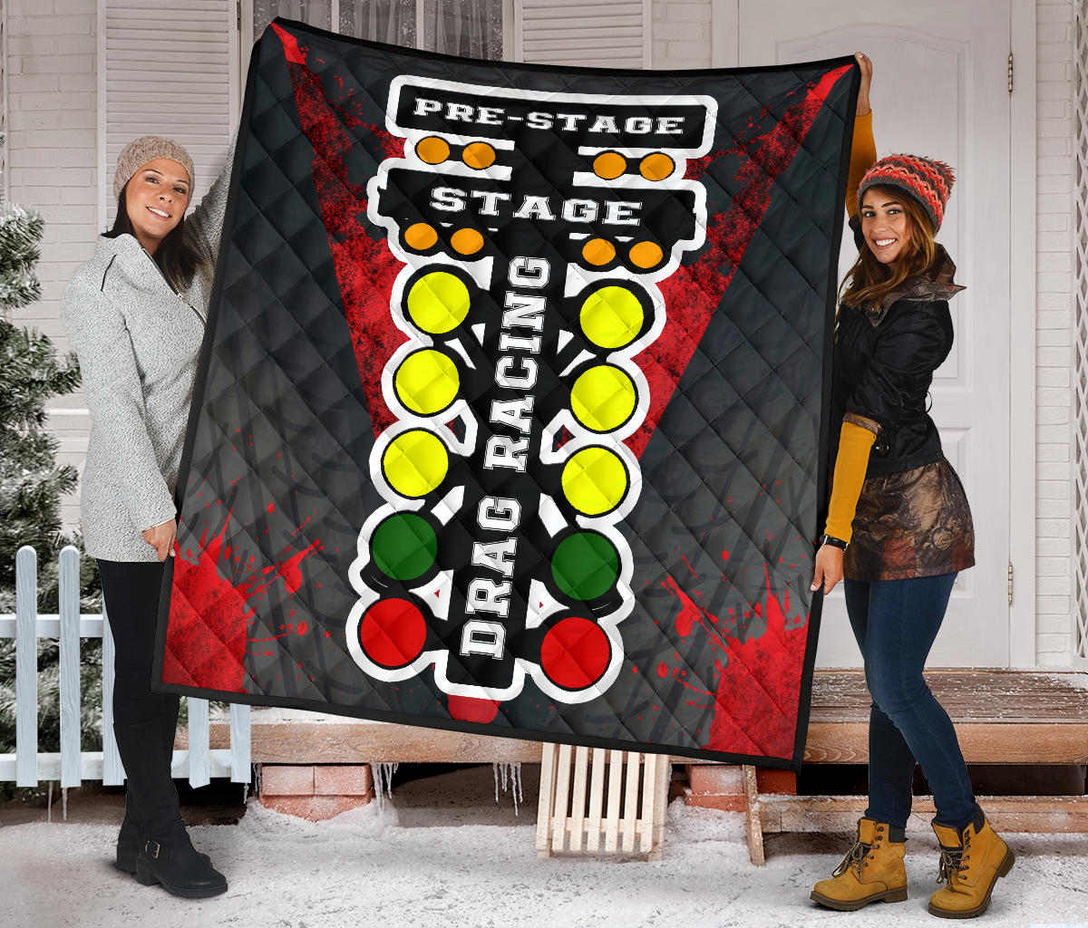 Two women displaying the red drag racing quilt outside a snowy porch, with bold staging lettering and colorful starting lights.