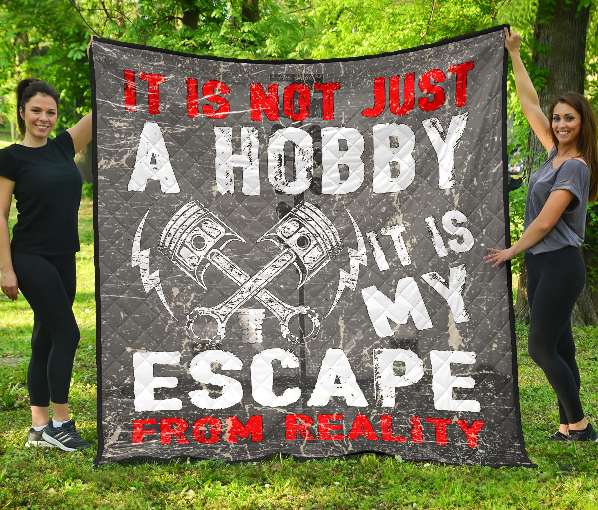 Two women holding a gray drag racing quilt outdoors, with crossed pistons and “It is not just a hobby, it is my escape from reality” lettering.